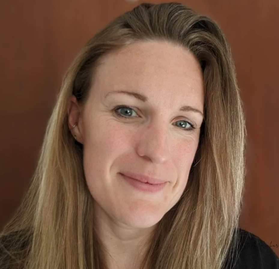 A woman with long, light brown hair and a slight smile stands in front of a wooden background. She is wearing a black top and a silver necklace.