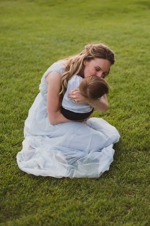 A black and white image of woman in a white dress kneeling on the grass, hugging and kissing her child.