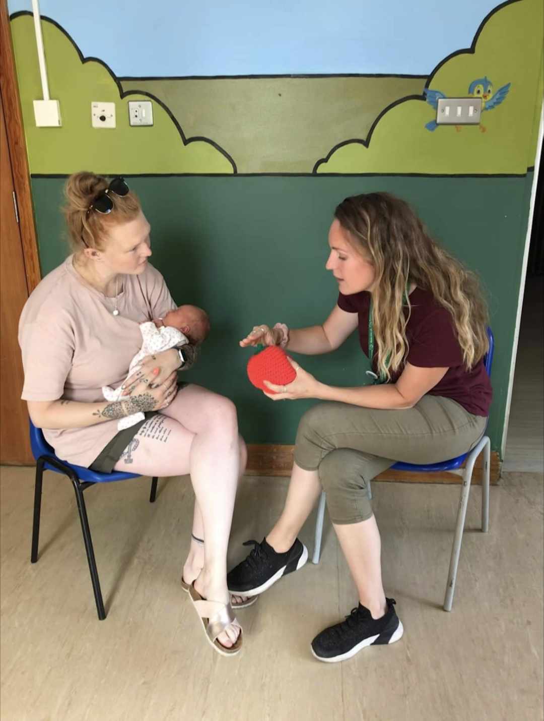 Two women in chairs, engaged in a warm conversation whilst one is caring for a baby nestled comfortably in her lap. The other woman is sitting with knitted breast and is showing the mother how to latch her baby, using her hands. 