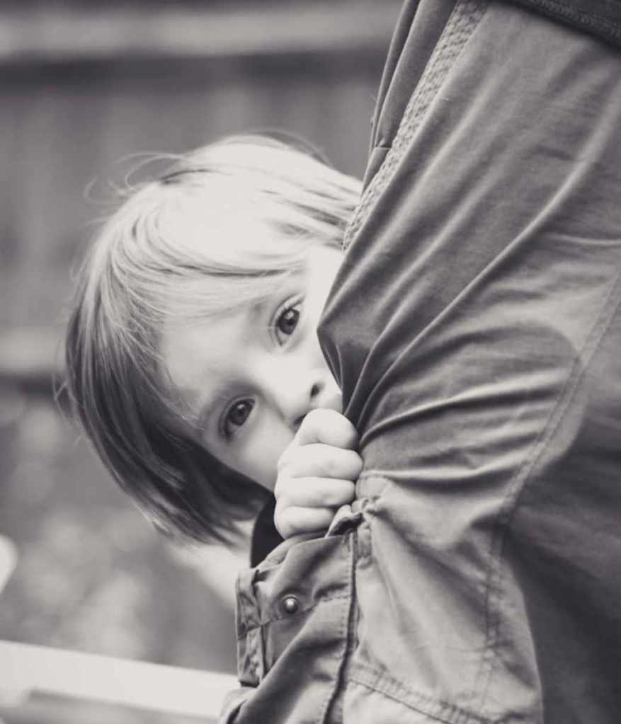 A child curiously peeks out from behind a man's leg, showcasing a moment of innocence and playfulness.