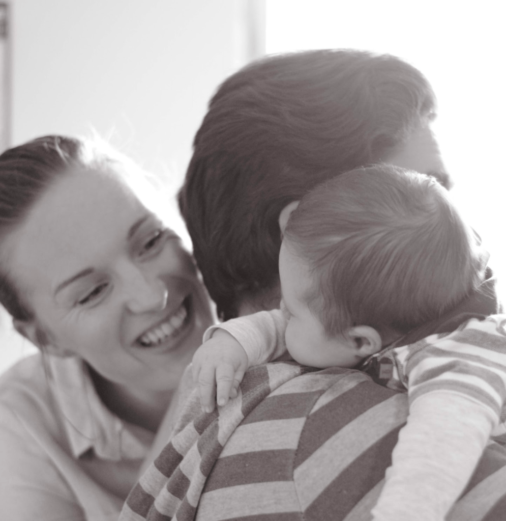 A monochrome image of a mother and father peeking at their new born baby. The baby is staring at the mother, whilst he lays over his father's shoulder